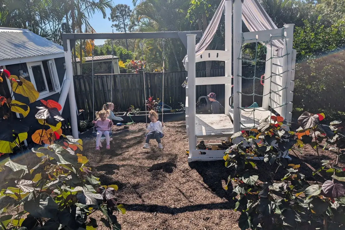 Children on swings in the backyard at Gizmo's, beside the cubby house and climbing frame.