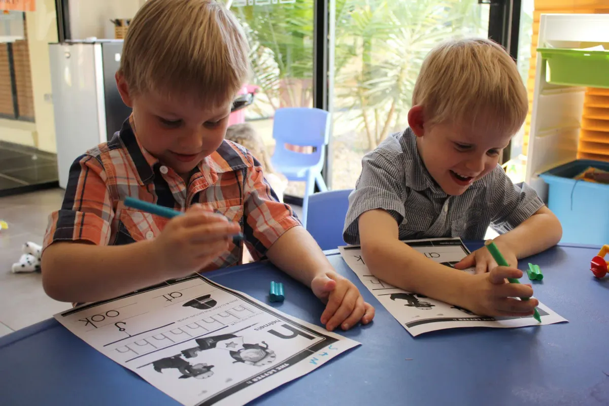 Two children at a table working through letter-sound worksheets with coloured markers.